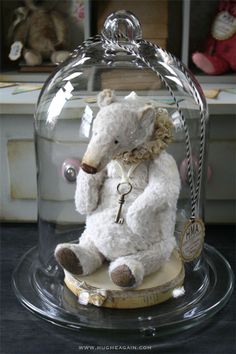 a white teddy bear under a glass clochet with a key in it's mouth