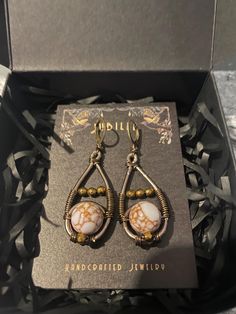 These earrings are handcrafted, featuring a unique design with a combination of materials. They are teardrop-shaped, created using metal wire wrapping that holds a central round stone with a distinctive marbled pattern. The stone is white with brownish-gold veins. There are additional small golden beads accenting the design, placed above and below the central stone within the teardrop frame. The earrings have lever-back closures for a secure fit. Copper Wire Wrapped Teardrop Earrings As Gift, White Metal Teardrop Earrings, White Teardrop Wire Wrapped Earrings, Wire Wrapped Metal Teardrop Earrings For Gift, Wire Wrapped Metal Teardrop Earrings As Gift, Copper Wire Teardrop Earrings, White Wire Wrapped Copper Wire Jewelry, Adjustable Nickel-free White Teardrop Earrings, White Brass Wire Wrapped Earrings