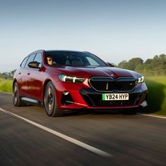 a red car driving down the road with trees in the backgrouund and grass on both sides