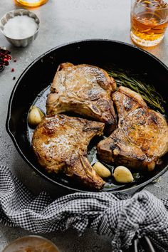 two steaks in a skillet with potatoes and herbs on the side, next to a glass of whiskey