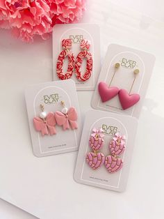 three pairs of pink and red earrings with heart shapes on them, sitting next to a pink flower