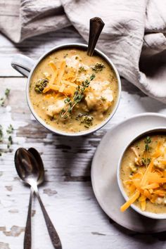 two bowls of soup with cheese and broccoli on the side next to spoons