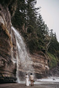 two people standing in front of a waterfall on the beach next to trees and water