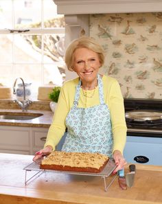 an older woman is holding a cake on a rack in the kitchen while smiling at the camera