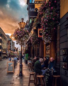 people sitting at tables on the sidewalk in front of buildings with flowers hanging over them