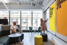 two people are sitting on couches in an office with large windows and yellow accents