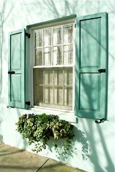 an open window with shutters on the side of a white wall and green planters below it