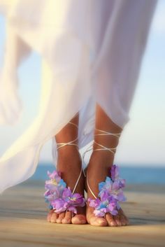 the feet of a woman wearing sandals with purple flowers on them and white dress underneath