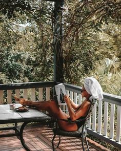 a woman sitting in a chair reading a book on a porch with trees behind her