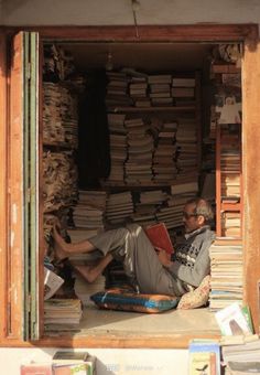 a man sitting in the doorway of a book store with stacks of books behind him