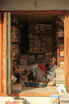 a man sitting in the doorway of a book store with stacks of books behind him