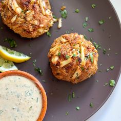 two crab cakes on a plate next to a bowl of dipping sauce and lemon wedges