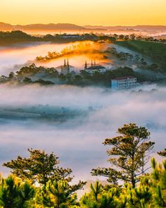 the sun is setting over some trees and buildings in the foggy valley with low lying clouds