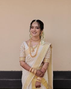 a woman wearing a white and yellow sari with gold jewelry on her neck, standing in front of a tan wall