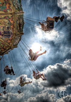 some people are riding on swings in the sky with clouds and sun shining through them