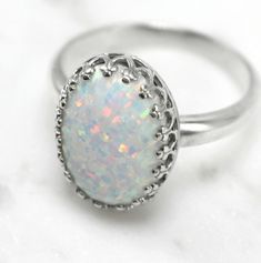 "This gorgeous white pinfire opal sits in a sterling silver 14 x 10mm (9/16\" x 3/8\") crown ring bezel that comes in 6, 7, 8, and 9 US ring sizes. The opal is lab-created with tones of sparkle in shades of pink, blue, lavender and green. Please read more about lab-created opals below. Opals are the birthstone of October but they are also known as the gem of choice by most and loved by many. If you have any questions please do not hesitate to contact me. What is a lab-created opal... Lab-created Oval Cabochon White Gold Opal Ring, Oval Cabochon Opal Ring In White Gold, Oval White Gold Opal Cabochon Ring, Classic White Opal Ring With Birthstone, Classic White Opal Birthstone Ring, White Opal Ring As A Gift, White Opal Ring For A Gift, White Opal Oval Cabochon Ring, Opal Birthstone Ring Oval Cabochon