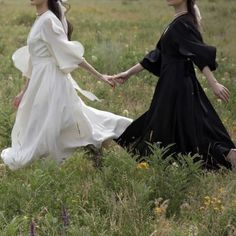 two women dressed in black and white walking through tall grass holding hands with each other