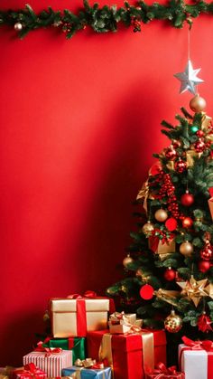 a christmas tree with presents under it in front of a red wall that has gold and silver ornaments on it