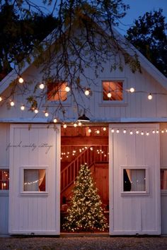 a christmas tree is lit up in front of a white barn with lights on it