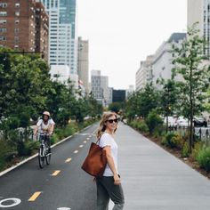 a woman standing on the side of a road with her bike in the background and another bicyclist riding down the street