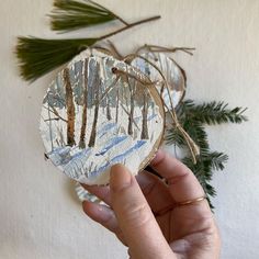 a hand holding a piece of paper with trees on it and pine branches in the background