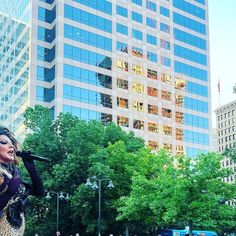 a woman is performing on stage in front of tall buildings with trees and people walking down the street