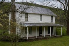 an old white house with a metal roof in the middle of a grassy field next to trees