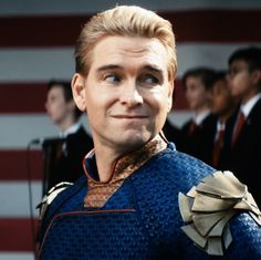 a man with blonde hair and blue shirt in front of an american flag, looking at the camera