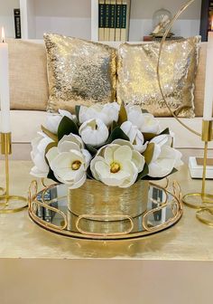 white flowers in a gold vase on a glass tray with candles and pillows behind it