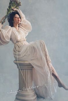 a woman sitting on top of a white pillar holding a flower in her hand and wearing a dress