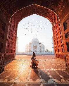 a woman sitting on the ground in front of an archway with birds flying around her