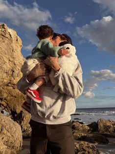 a man holding a child on the beach with rocks in the background and clouds in the sky