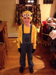 a young boy dressed up as a minion with goggles and overalls standing in a living room