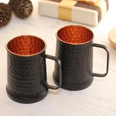 two black coffee mugs sitting on top of a white table next to pine cones