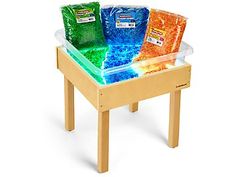 three bags of colored sand sit on a wooden table with clear plastic trays in front of them