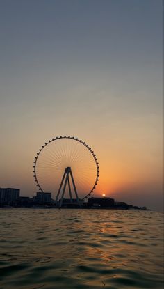 the sun is setting behind a ferris wheel on the water in front of some buildings