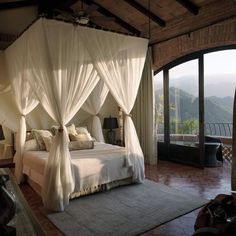 a bed with white drapes and pillows in a bedroom next to a large window