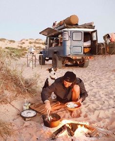 a man sitting in the sand next to a campfire with his dog looking on