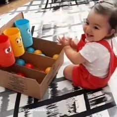 a baby sitting on the floor next to a cardboard box with cups in it and smiling