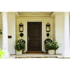 two planters with white flowers are on the front porch of a house, next to a brown door