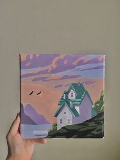 a hand holding up a small painting with a house on the hill in the background