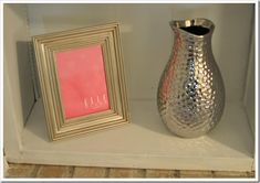 a silver vase sitting on top of a white shelf next to a framed picture and a pink card