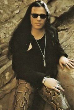 a man with long hair and sunglasses sitting on a rock next to a large snake