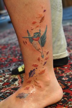 a hummingbird tattoo on the foot of a woman's right leg, with leaves falling from it