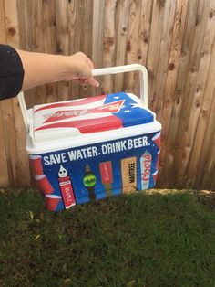 a cooler with drinks in it sitting on the grass next to a fence and hand