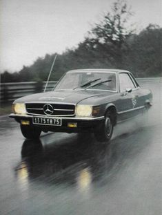 an old mercedes benz is driving down the road on a rainy day with trees in the background