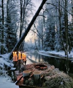 candles are lit on a table in the middle of a snowy forest by a river