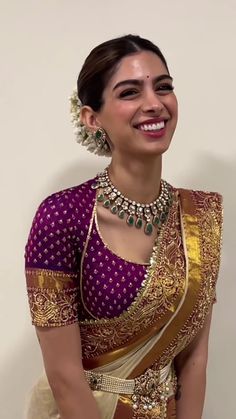 a woman in a purple and gold saree smiles at the camera while standing next to a wall