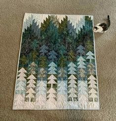 a cat is sitting on the floor next to a quilt that has trees in it