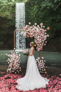 a woman standing in front of a waterfall with pink flowers around her and holding a bouquet